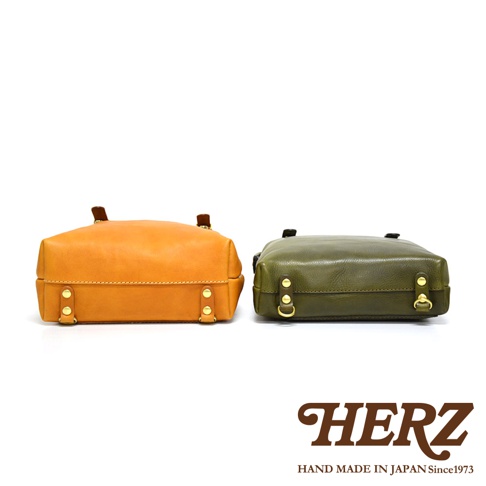 職人手作皮革後背包 R-143，展示卡其色與墨綠色兩款。皮質背包側面有金屬扣環與鉚釘裝飾，設計簡潔。