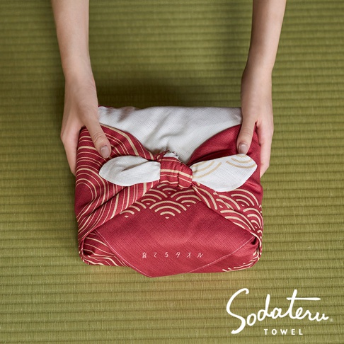 一雙手拿著用紅色和白色水波紋圖案的風呂敷包起來的毛巾，下方有 Sodateru TOWEL 的品牌標誌。