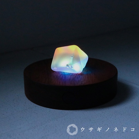 合光稜鏡石，多面體造型，透明材質，內部散發彩虹色光芒，置於圓形木製底座上，整體氛圍柔和。