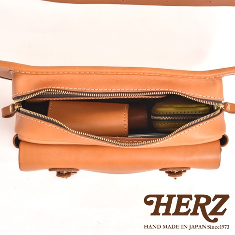 職人手作皮革騎士隨身斜背包，淺棕色牛皮材質，主隔層拉鍊打開，內有棕色皮製捲筒狀小物、綠色皮製零錢包，以及其他物品。