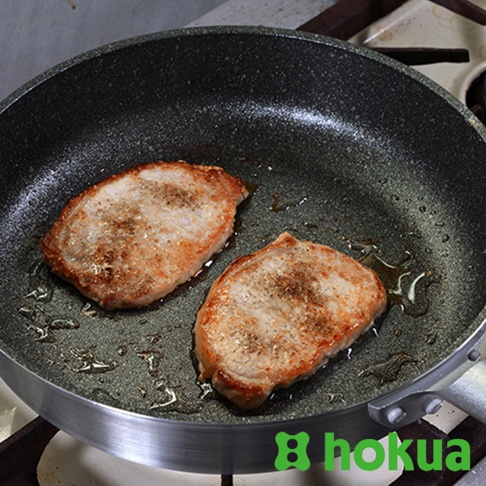 一個30公分的黑色不沾炒鍋，鍋內煎著兩塊金黃色的豬排，豬排表面撒有胡椒粉，鍋底有少量油。