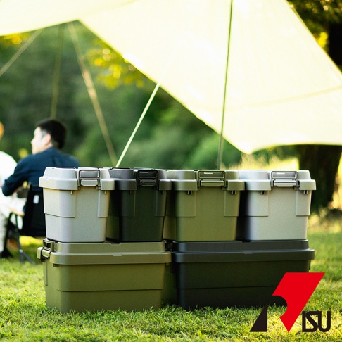 堆疊的 TRUNK CARGO 可堆疊多功能耐重收納箱，共三色：卡其色、深綠色和灰色。箱體由堅固的塑料製成，帶有兩個鎖扣和一個把手。產品放置在戶外的草地上，背景是模糊的樹木和黃色的遮陽篷。