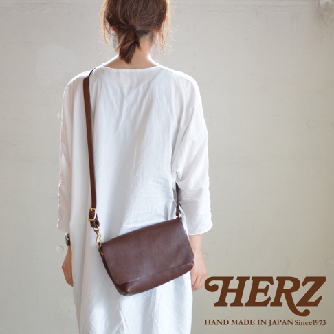 一名女子背對鏡頭，身穿白色寬鬆上衣，斜挎一個棕色皮革翻蓋斜背包，包包上有金屬扣環，右下角有HERZ品牌標誌和Hand Made in Japan Since 1973字樣。
