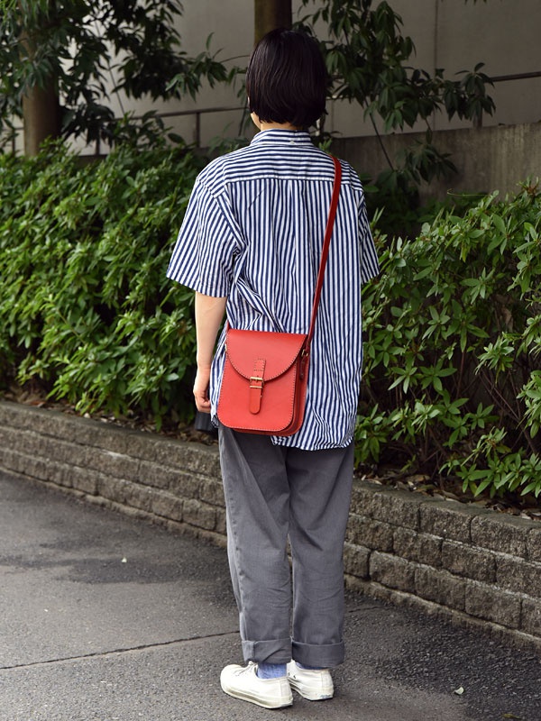 紅色皮革翻蓋迷你單肩包，配有金色搭扣和細長紅色肩帶，斜挎於身穿藍白條紋襯衫和灰色長褲的人身上。