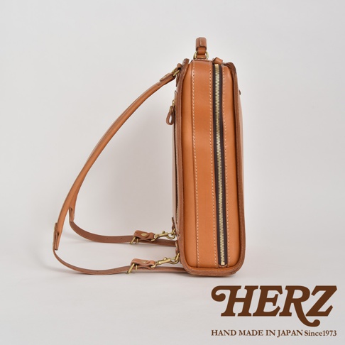 職人手作皮革商務電腦後背包 R-25，採用鞣製皮革材質，呈現溫潤的棕褐色調。背包側面可見拉鍊細節與可調節的皮革背帶，整體線條簡潔，適合商務場合使用。