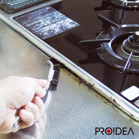 一隻手握著 J 型隙縫刷，正在清潔爐灶邊緣的油垢，刷毛為黑色，刷柄為透明塑膠。