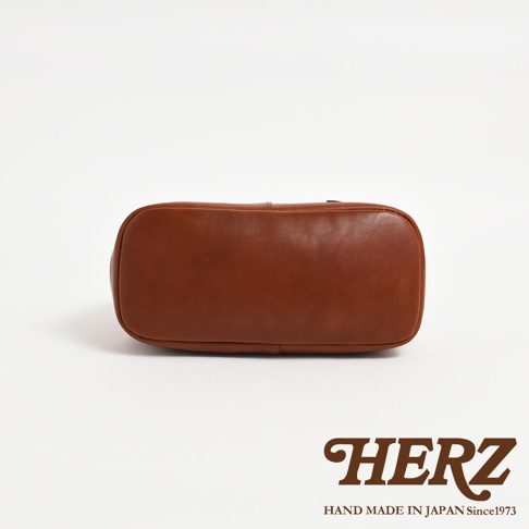 HERZ 品牌標誌，下方是 HAND MADE IN JAPAN Since1973 字樣，前景為日本 HERZ 品牌褐色皮革托特包底部特寫。