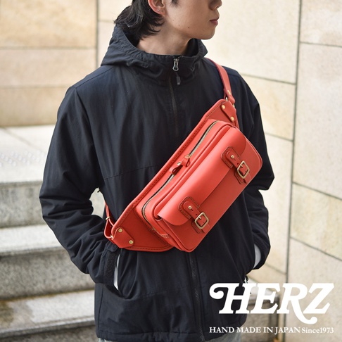 職人手作皮革騎士隨身斜背包，採用鮮豔的紅色皮革製成，包款具有雙層拉鍊主袋、前方翻蓋口袋，並以銅釦和皮革繫帶固定，斜背於男性模特兒身上。