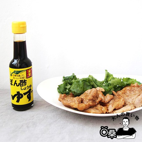 一瓶金色的玻璃瓶裝醬油，瓶身黃色標籤印有日文，瓶蓋為黃色。旁邊是一盤白色盤子，盛有綠色蔬菜和幾塊棕色的肉。