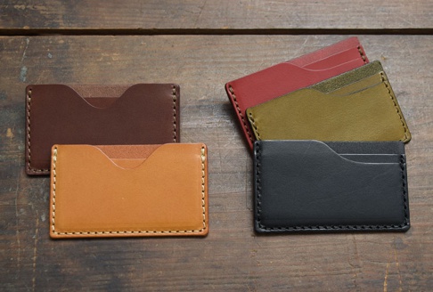五款皮革卡片夾，顏色包含深棕、淺棕、紅、橄欖綠、黑色，以手工縫線製成，樣式簡約，擺放在木質背景上。