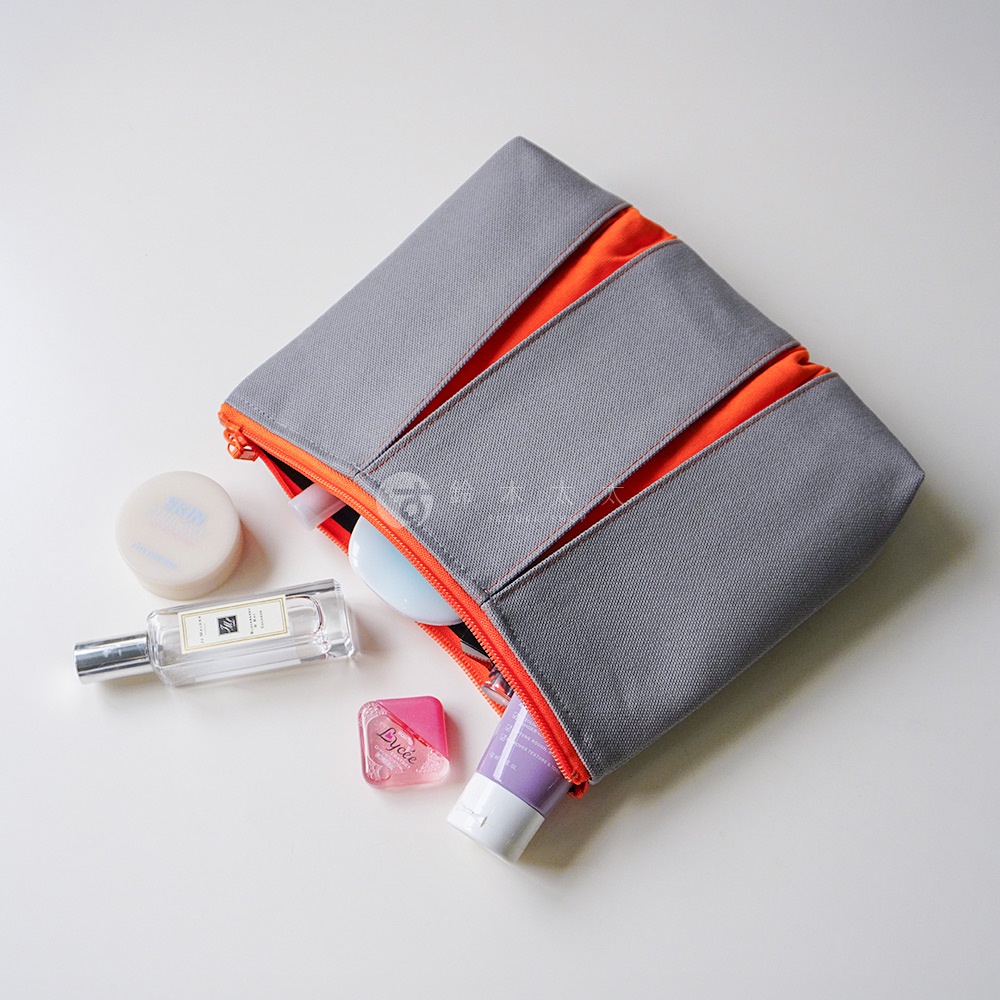 一個灰色帆布材質的長方形收納包，內部飾以鮮豔的橘色內襯與拉鍊，呈現半開啟狀態。包包周圍散落著護膚霜圓罐、香水瓶、粉色小型眼藥水以及紫色軟管狀美妝用品，整體採俯視角度拍攝，展現出簡約且具現代感的收納空間。