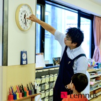 圓形木質邊框時鐘，白色錶盤，黑色數字和刻度，紅色時針和藍色分針。右側是一位身穿藍色圍裙的男子，正伸手調整時鐘。左側牆上掛著一個藍色的小鬧鐘，下方擺放著彩色鉛筆。