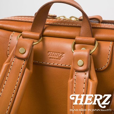 職人手作皮革軟皮商務後背包 R-28，近距離特寫，展現棕褐色光滑皮革材質、精緻的縫線細節、黃銅色五金扣環，以及壓印的「HERZ」品牌標誌和「MADE IN JAPAN」字樣。