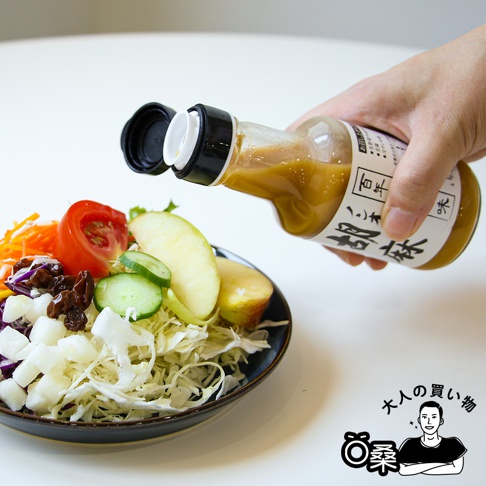 一隻手拿著透明玻璃瓶，瓶內裝有淺棕色的芝麻沙拉醬，瓶身有日文標籤。沙拉醬正從瓶口滴入裝有鮮豔蔬菜和水果的藍色碗中，碗內有高麗菜絲、番茄、小黃瓜片、蘋果片、胡蘿蔔絲和葡萄乾。