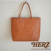 一個 tan 色皮革托特包，有雙提把，包包正面有縫線細節，底部中央印有 HERZ 商標和 HAND MADE IN JAPAN Since1973 字樣