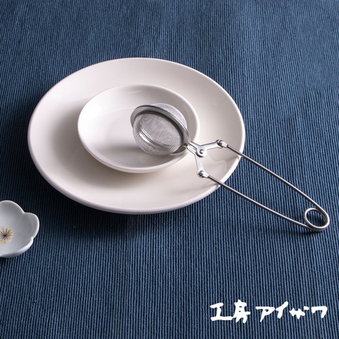 銀色不鏽鋼製的泡茶器，帶有網狀濾網和長柄夾子，放置於白色盤子上，背景為藍色紋理布墊。