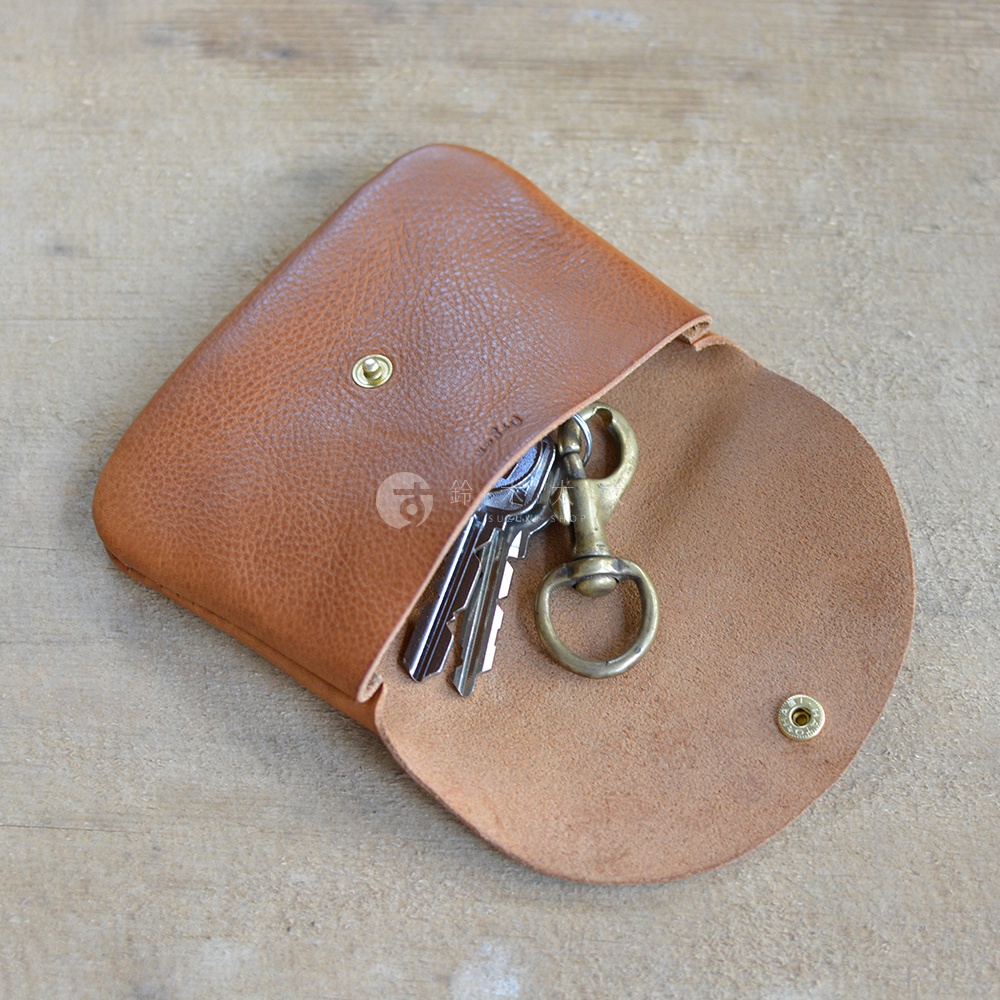 淺棕色皮革製成，帶有鈕扣閉合的職人手作小物收納袋，內有鑰匙圈及數把鑰匙。