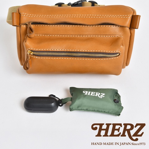 職人手作皮革兩用腰掛斜背包，採用棕褐色皮革製成，配有黃銅色拉鍊和米色織帶。包款具有多個拉鍊隔層，並展示了黑色耳機盒和綠色收納袋。