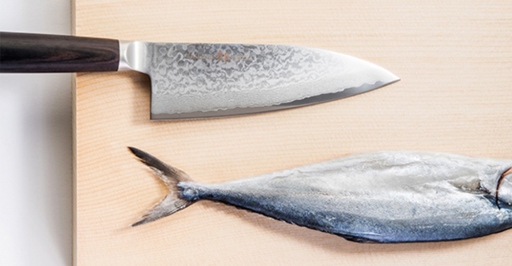 帶有黑色木柄的日式Deba魚肉料理刀，刀刃呈現獨特的大馬士革紋路，與一條橫躺在淺色木砧板上的銀灰色魚相鄰。