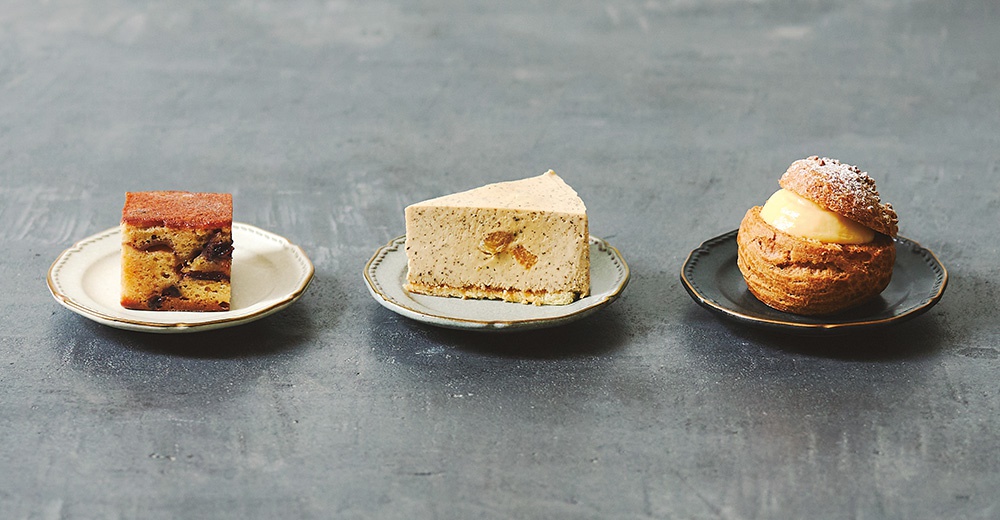 三塊不同口味的蛋糕，分別是果乾蛋糕、慕斯蛋糕和泡芙，擺放在盤中，背景為灰色紋理。