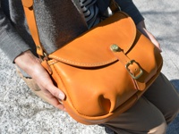 一個橙棕色皮革製成的馬鞍包，配有可調節的皮帶和黃銅扣環，由一位女士斜挎著。