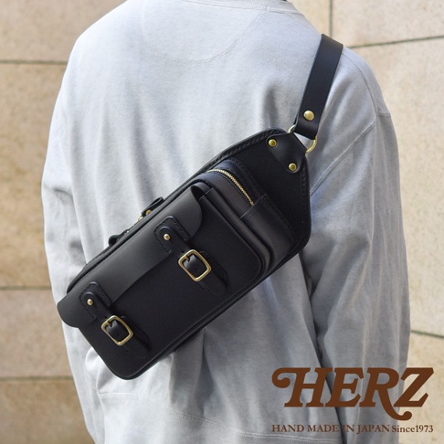 黑色皮革騎士斜背包，採用職人手作工藝，配有黃銅釦環、拉鍊和铆釘，展現復古風格。