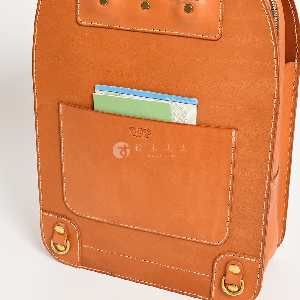 職人手作皮革商務休閒兩用後背包 R-36，展示為焦糖棕色皮革，設有一個前置口袋，內有綠色筆記本和地圖，口袋上方有「HERZ」和「鈴木太太」品牌標誌，兩側下方有黃銅色D型環。