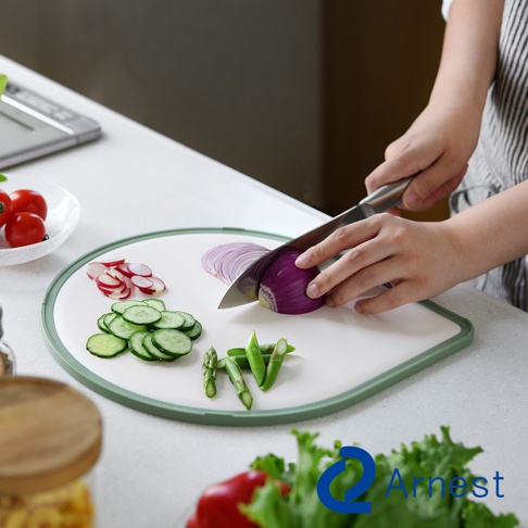 淚滴型抗菌料理砧板，白色光滑表面，周圍有淺綠色邊框。砧板上擺放著切好的小黃瓜片、紫洋蔥、小番茄和四季豆，一位女士正用刀切著紫洋蔥。