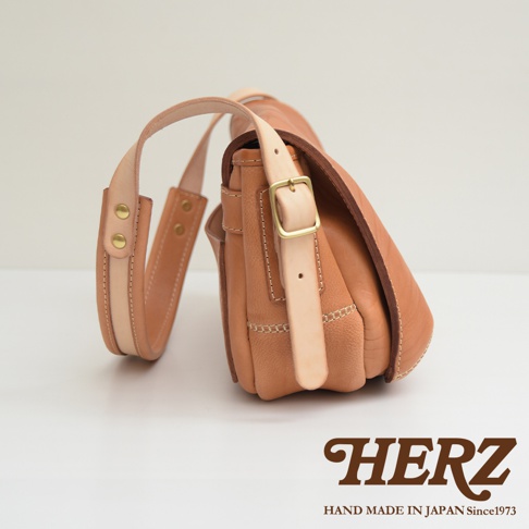 職人手作皮革郵差包，淺棕色，帶有金色扣環和可調節肩帶，皮革質感，縫線明顯。