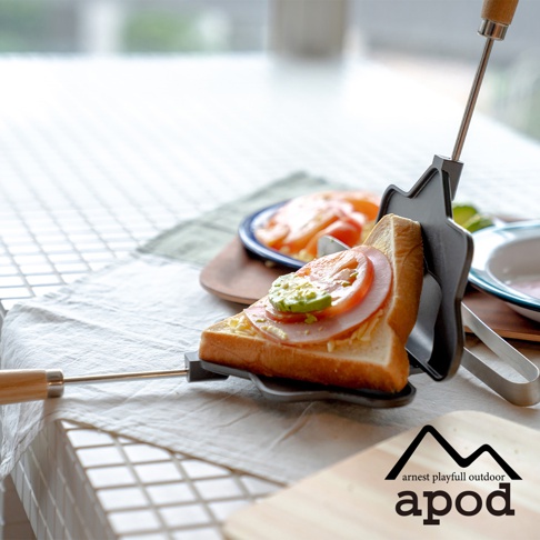 一個帶有木質手柄的黑色星形熱壓三明治烤夾，夾著一片塗有芥末醬、火腿、番茄和酪梨的三明治。背景是淺色的桌面和一塊木質砧板。