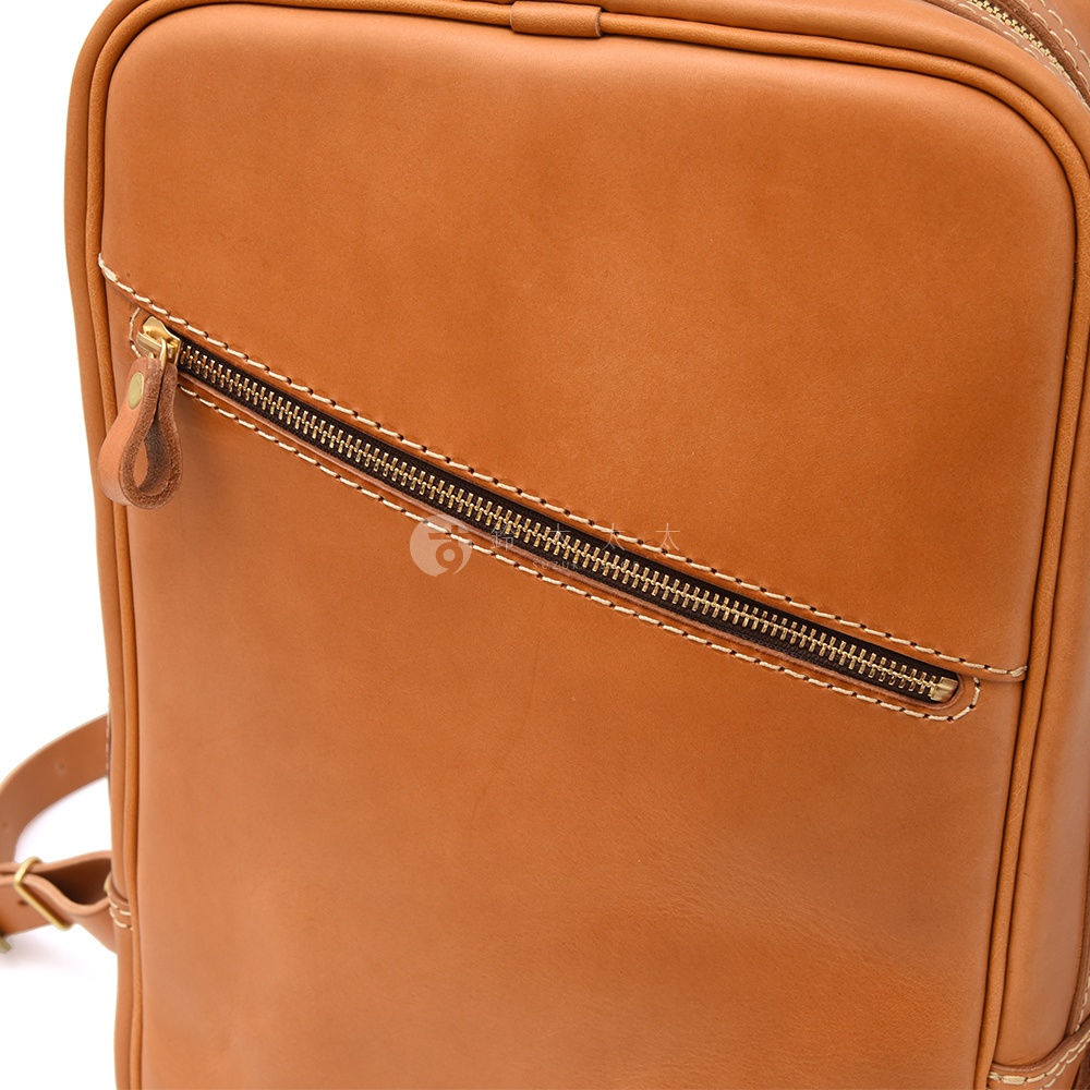 棕褐色皮革長方型後背包，表面有一個斜向的拉鍊口袋，附有皮革拉環。車縫線明顯，材質呈現自然光澤。