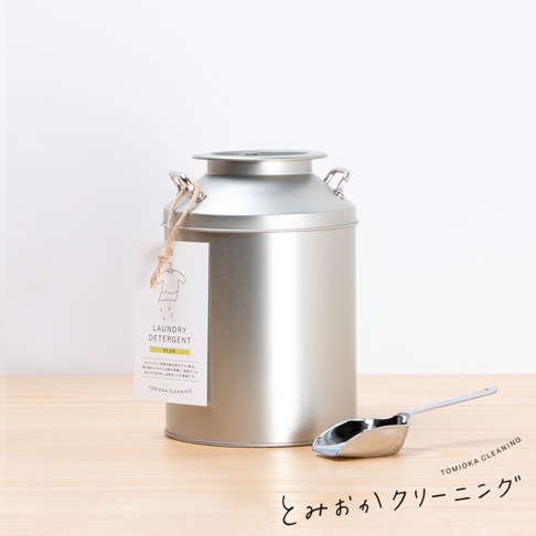 銀色金屬牛奶罐造型洗衣粉罐，附有吊牌標示「LAUNDRY DETERGENT PLUS」，旁有一銀色金屬量勺。