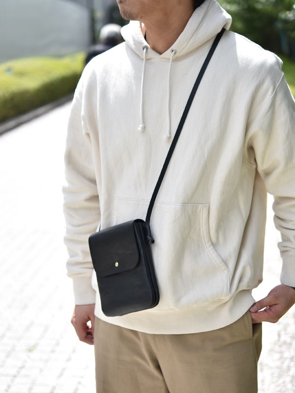 黑色皮革製直式隨行袋，帶有金色鈕扣閉合。袋子斜挎在穿著米白色連帽衫和卡其色褲子的男性身上。