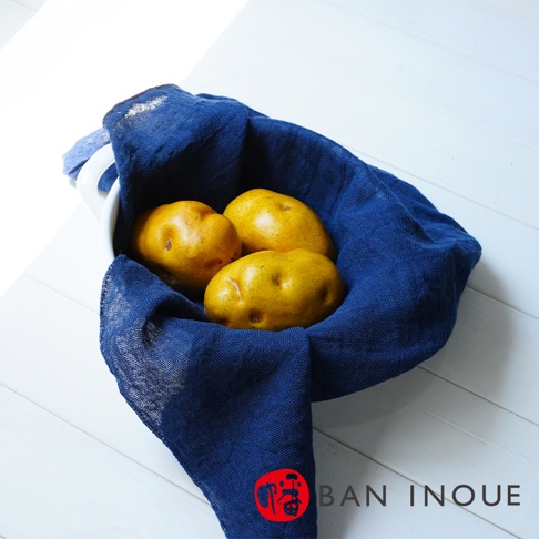 BAN INOUE 品牌字樣標誌於藍色布巾下方的紅色印章旁，畫面中有三顆黃色馬鈴薯被藍色布巾包覆著，置於白色碗中，背景為白色木紋桌面。