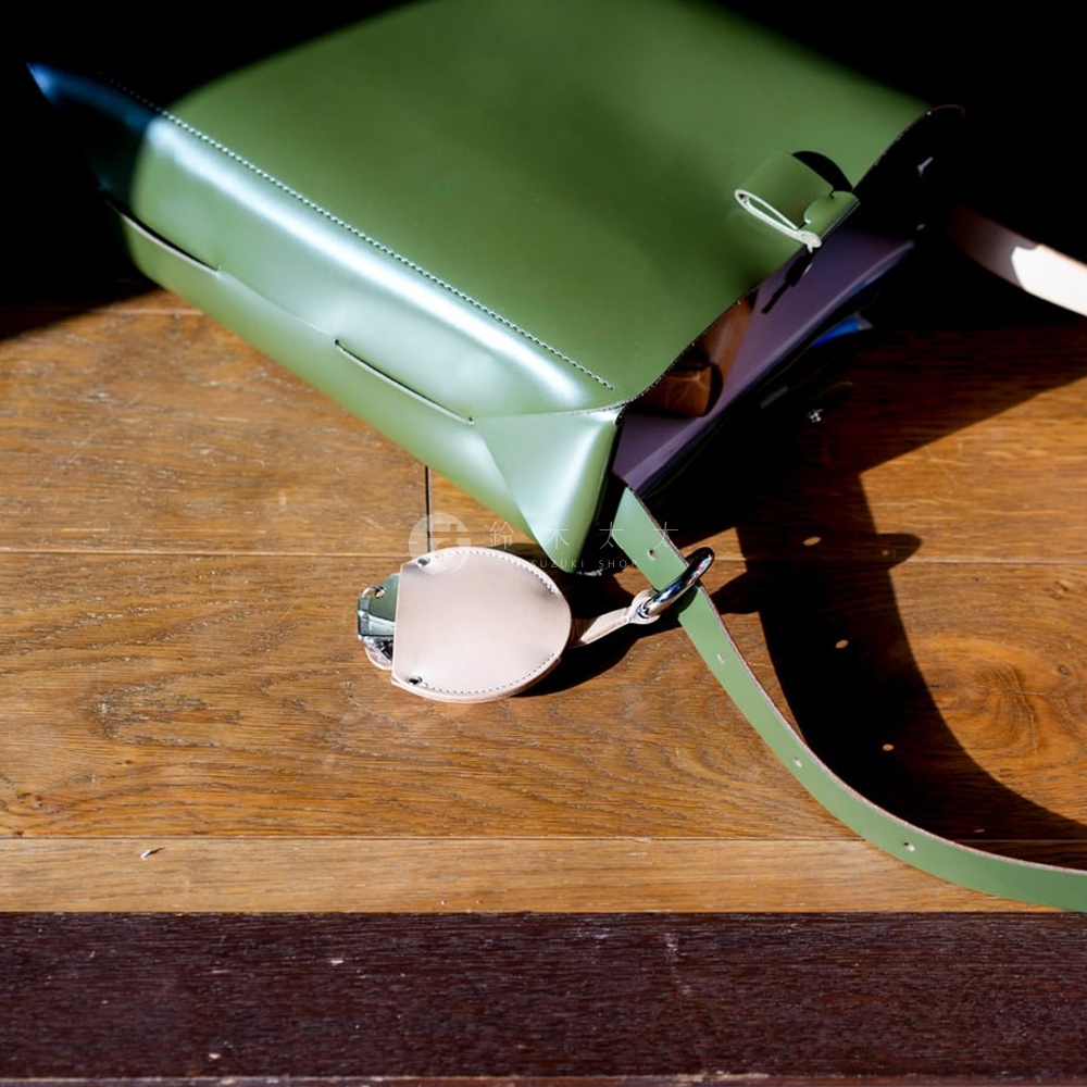 綠色皮質包斜掛在木質桌面上，一旁垂掛著一個米白色皮革製成的圓形鑰匙圈，上面有金屬扣環。