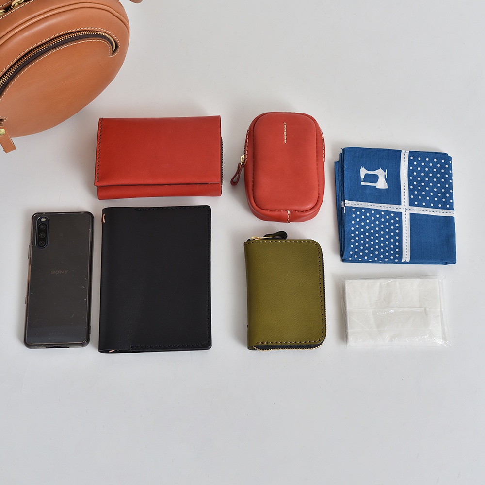 皮革小圓包 CK-19 S (共5色) 的內容物展示：一個橘棕色皮革小圓包、一個紅色短夾、一個紅色拉鍊小包、一個橄欖綠拉鍊零錢包、一支黑色手機、一本黑色筆記本、一塊藍色印花手帕，以及一包白色面紙。