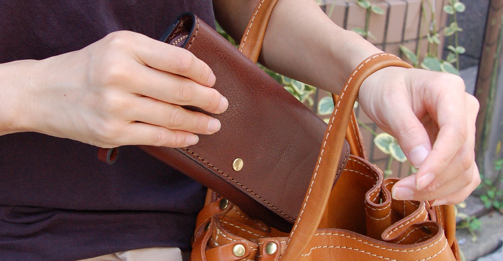 一隻手正將一個深棕色皮革長夾放入一個淺棕色皮革托特包中。長夾上有縫線和一個金色金屬鈕扣。