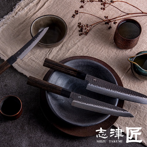 三把「玄 / Chef's knife 主廚刀18cm」，刀刃呈現鍛造紋理，握柄為深棕色木質。兩把刀置於黑色圓盤中，另一把刀斜放在米白色布料上，旁有小茶杯與綠色陶製醬料碟。