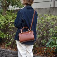 一名女子身揹一個棕色皮革迷你波士頓包，包款配有提把及可拆卸肩帶，側邊與底部有金屬腳釘裝飾，整體呈現經典復古風格。