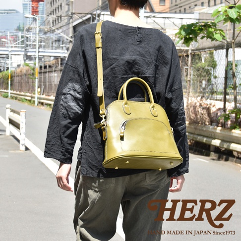 橄欖綠皮革手提包，貝殼造型，配有可拆卸肩帶，正面設有拉鍊口袋，包底有四個金屬腳釘，由職人手工製作。