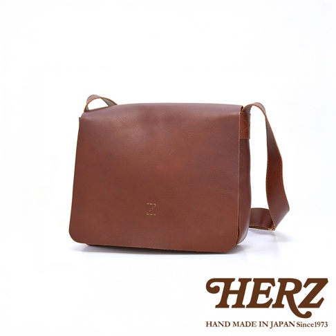 一個棕色的皮革斜背包，正面壓印有「Z」字樣，下方有「HERZ HAND MADE IN JAPAN Since1973」的品牌標誌。