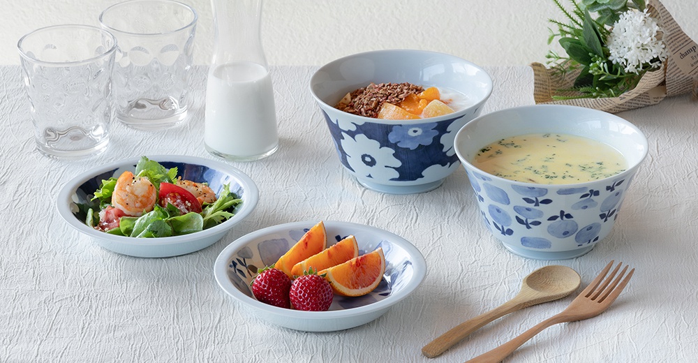 白色陶瓷丼飯碗，碗身繪有藍色花紋，碗內盛有沙拉、水果麥片或湯，搭配透明玻璃杯與木質餐具。