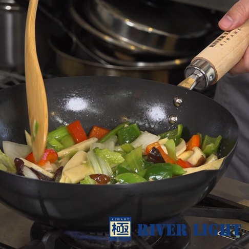 黑色不易生鏽炒鍋中，烹調著紅、綠甜椒、洋蔥、香菇等什錦蔬菜，木製鍋柄清晰可見，鍋身有兩個銀色鉚釘。