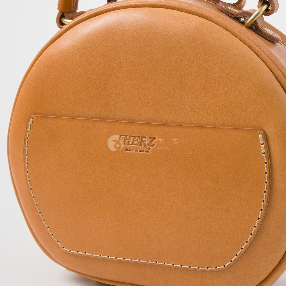 日本HERZ職人手作皮革圓形包，近拍皮革材質與精緻縫線細節，中央壓印有HERZ品牌標誌及MADE IN JAPAN字樣。