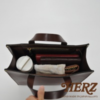 深棕色皮革直式托特包，內部裝有手機、皮夾、鑰匙包及一個白色圓形物品。包包採用職人手作皮革材質，做工精緻，展現高品質。