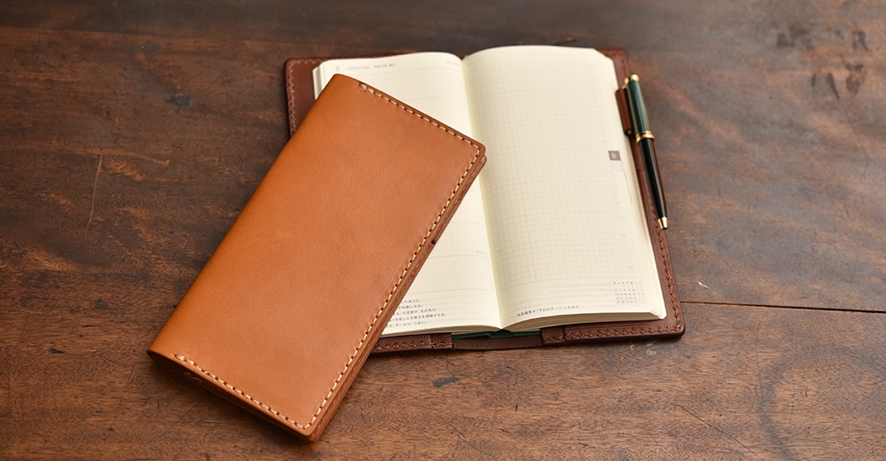 職人手作皮革旅行記事本書套，採用淺棕色皮革，搭配白色車縫線，內含一本網格頁面的筆記本及一支黑色鋼筆。