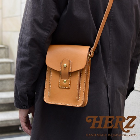 職人手作皮革休閒散步包，淺棕色，方形設計，翻蓋開合，附有金屬釦和皮製繫帶，側邊有縫線裝飾，斜背於身。