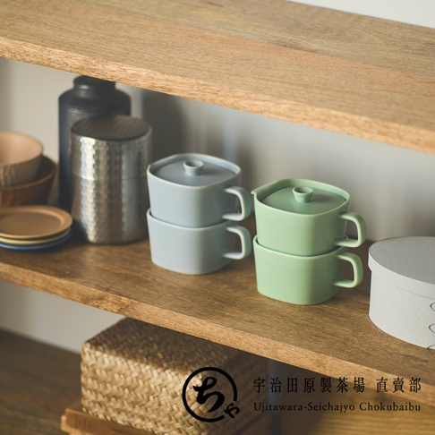 木質層架上擺放著精緻的居家用品，中央疊放著兩組美濃燒一人用馬克杯，左側為淡藍色，右側為淺綠色，杯身設計簡約並配有同色杯蓋。旁側還有金屬茶葉罐、碗盤與編織置物籃，畫面下方標註著宇治田原製茶場直賣部的品牌資訊，營造出溫馨舒心的居家茶飲氛圍。