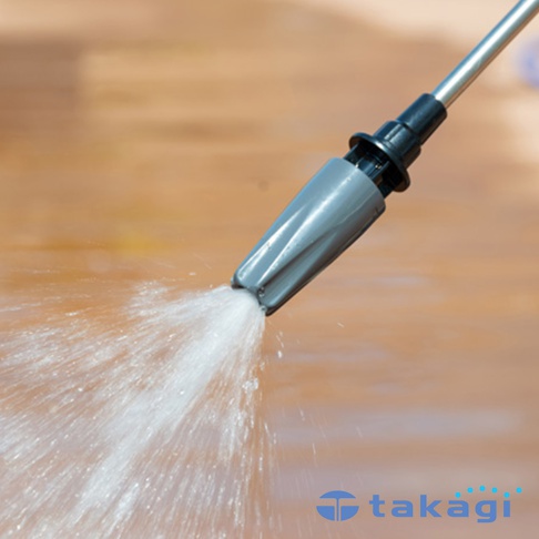 長型洗車清潔噴水槍的灰色噴頭，正在噴射強勁的水柱，水花四濺，背景為淺棕色的木質地板。