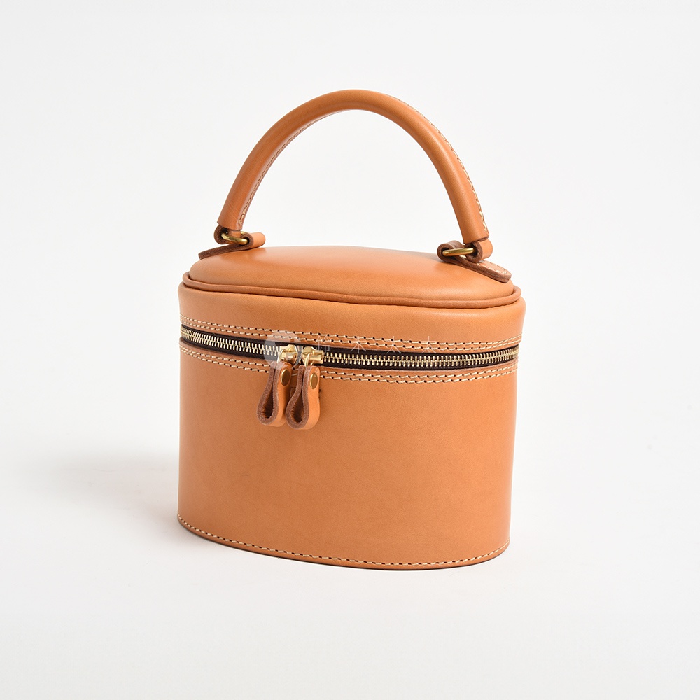 天然皮革製成的圓筒形化妝手提包，帶有雙拉鍊和頂部提把，呈現自然的棕褐色。